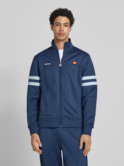 Männermodel trägt Sweatjacke aus Baumwoll-Mix Modell 'ROMA' von Ellesse in Dunkelblau mit weißen Streifen, Gerade Passform, Stehkragen, durchgehender Reißverschluss, Label-Details, Eingrifftaschen und elastische Abschlüsse, Rückenlänge 70 cm, Ärmellänge 68 cm, Ganzkörperansicht von vorne.