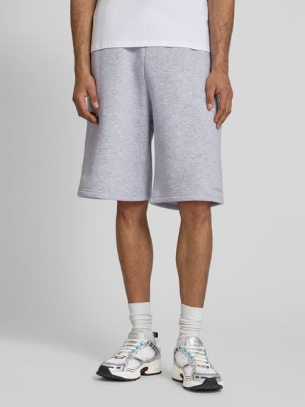 Mannenmodel draagt Sweatshorts met Logo-Print van REVIEW in lichtgrijs, katoenmix, loose fit, elastische band, logoprint, binnenbeenlengte 28 cm, bandwijdte 45 cm, detailopname onderlichaam.