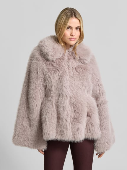 Frauenmodel trägt Flared Jacke aus Kunstfell von ROTATE in hellem Flieder, Trompetenärmel, gerade Passform, Kunstfell-Textur, Nahaufnahme Oberkörper. Innenfutter mit Viskose-Anteil, Rückenlänge 60 cm, Brustweite 39 cm, Ärmellänge 66 cm.