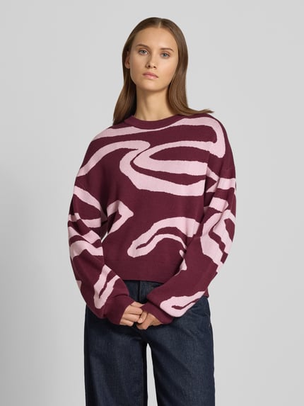 Frauenmodel trägt Relaxed Fit Pullover aus Viskose-Mix Modell 'Charlott' von NOISY MAY® mit Animal-Print in Bordeaux und Rosa, gerippte Abschlüsse, Rundhalsausschnitt, Langarm, Ganzkörperansicht von vorne.