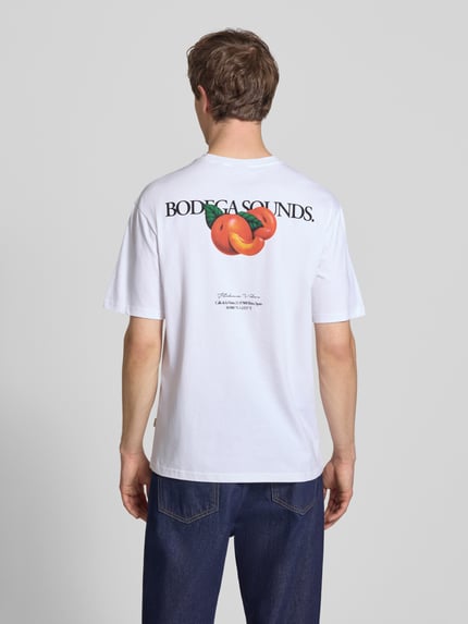 Männermodel trägt Relaxed Fit T-Shirt aus reiner Baumwolle Modell 'TASTY' von Only & Sons, weiß, Rückseite mit Schriftzug-Print und Rücken-Print, Rundhals, kurzarm, Ganzkörperansicht Rückansicht.
