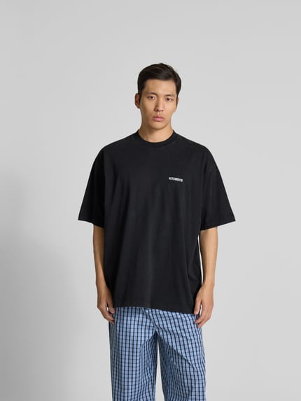 Männermodel trägt Relaxed Fit T-Shirt aus reiner Baumwolle von VETEMENTS in Schwarz mit Rundhalsausschnitt und Label-Stitching, Ganzkörperansicht von vorne.