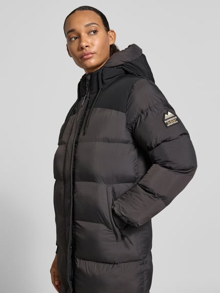 Frauenmodel trägt Steppmantel mit Kapuze und verdecktem Reißverschluss von Superdry in Dunkelgrau und Schwarz, sichtbare Eingrifftaschen, strukturierte Oberfläche, Regular Fit, Ganzkörperansicht von vorne. Rückenlänge 108 cm, Schulterbreite 42 cm, Brustweite 53 cm, Ärmellänge 61 cm.