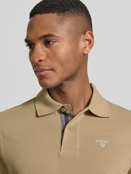 Männermodel trägt Regular Fit Poloshirt aus reiner Baumwolle von Barbour in Optik: Unifarben mit Label-Stitching-Applikation, Nahaufnahme Oberkörper.