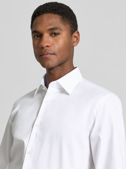 Męski model nosi Slim Fit Businesshemd aus reiner Baumwolle Modell 'HANK' von BOSS, biała popelina, długi rękaw, Kent kołnierzyk, jednorzędowa listwa guzikowa, sportowy mankiet, aplikacja z logo, wygląd jednolity, krój slim fit, widok z przodu zbliżenie na górną część ciała.