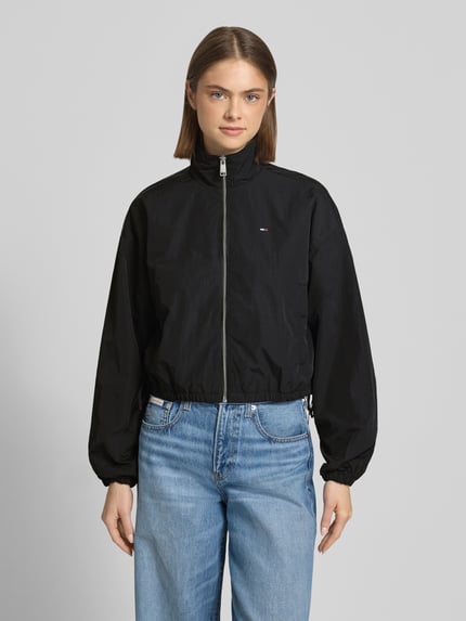 Frauenmodel trägt Relaxed Fit Blouson mit Taillenbändern von Tommy Jeans in Optik: Unifarben, Schwarz, Reißverschluss, Logo-Stitching, seitliche Eingrifftaschen mit Druckknopf, Stehkragen, Langarm, elastischer Ärmelabschluss, glatte Struktur, Ganzkörperansicht von vorne.