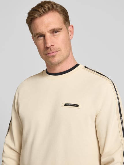Męski model nosi Sweatshirt mit Label-Details und Rundhalsausschnitt od EA7 Emporio Armani, mieszanka bawełny, krój regular fit, jasny kolor, detale z logo, okrągły dekolt, długość tyłu w rozmiarze M: 63 cm, długość rękawów w rozmiarze M: 66 cm, zbliżenie na górną część ciała.