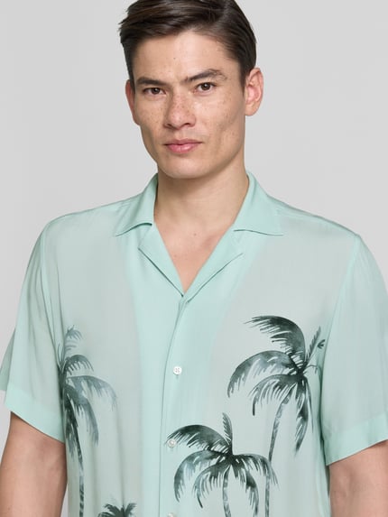 Mannenmodel draagt Regular Fit Freizeithemd aus Viskose Modell 'RAYER_M' van BOSS Orange, groen met palmbomen, resortkraag, kort mouw, all-over print, pure viscose, Nahaufnahme Oberkörper.