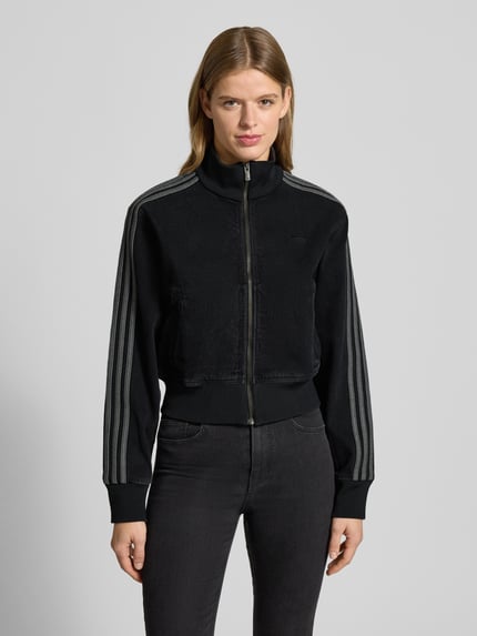 Kobiece modelka nosi kurtkę z zamkiem błyskawicznym od adidas Originals, krój regular fit, skrócony krój, prążkowane wykończenia, stójka, wpuszczane kieszenie, wyhaftowane logo, długość tyłu 51 cm, szerokość ramion 45 cm, szerokość klatki piersiowej 41 cm, długość rękawów 62 cm, kolor czarny, gładka tekstura, całkowity widok z przodu.