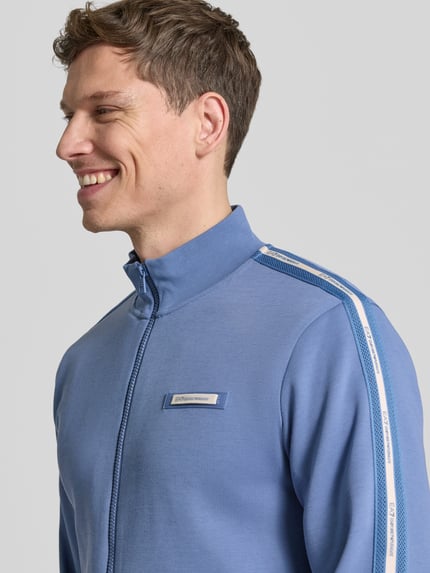 Männermodel trägt Sweatjacke aus Baumwoll-Mix von EA7 Emporio Armani in Blau mit Galonstreifen, Label-Detail, Nahaufnahme Oberkörper.