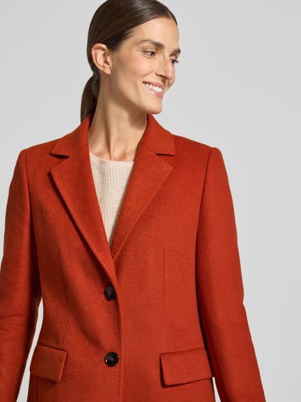 Modelka kobieca nosi krótki płaszcz z Pattentaschen von JAKE*S WOMAN COLLECTION, krój regular fit, jednokolorowy, długi, detal z logo, szwy działowe, guziki, kołnierz z połami, rozporek, zaszewki, zapięcie na 2 guziki, długi rękaw, kieszenie z patką, zbliżenie na górną część ciała.