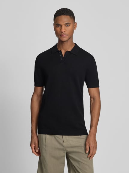 Männermodel trägt schwarzes Slim-Fit Poloshirt Modell 'Triton' von Drykorn aus Reine Baumwolle mit Polokragen und Strukturmuster, Kurzärmelig, Rückenlänge bei Größe M 67 cm, Ganzkörperansicht von vorne.