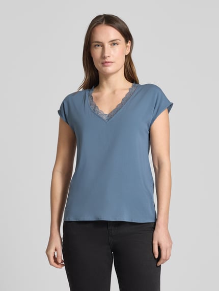 Modelka nosi Regular Fit T-Shirt mit Spitzenkragen Modell 'BELLA' von Vero Moda, unikatowy niebieski kolor, jersey-stretch, krój regular fit, V-dekolt z koronką, krótki rękaw, materiał wiskozowy, optyka jednolita, widok całej sylwetki z przodu.