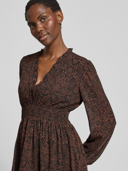 Frauenmodel trägt Minikleid mit elastischem Taillenband und V-Ausschnitt von Apricot aus Chiffon, taillierter Schnitt, Allover-Look, Rüschen, dunkelbraune Farbe mit filigranem Allover-Muster, Rückenlänge bei Größe S 85 cm, Brustweite 45 cm, Ärmellänge 67 cm, Nahaufnahme Oberkörper.