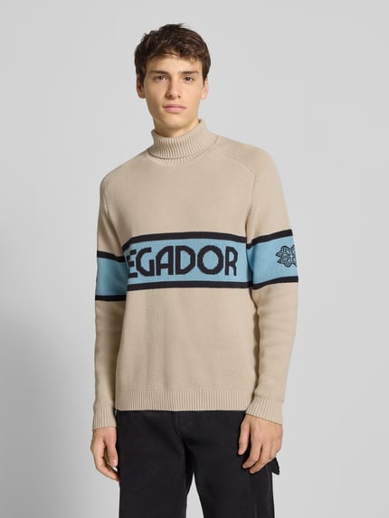 Mannenmodel draagt Strickpullover mit Rollkragen van Pegador in beige met blauw label-print, katoenmix, regular fit, ribboorden, vooraanzicht bovenlichaam.