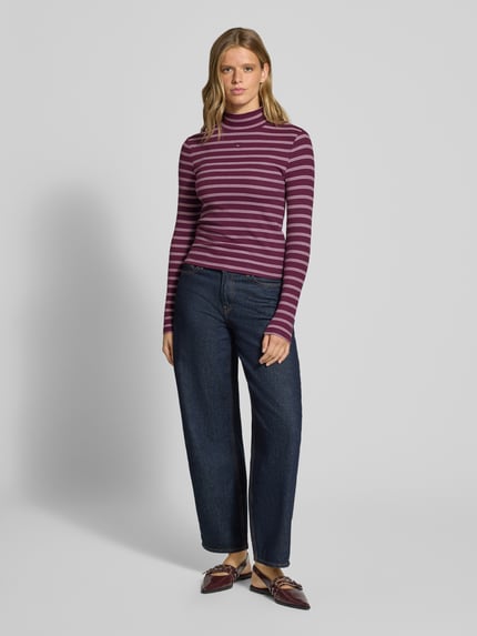 Frauenmodel trägt Slim Fit Langarmshirt aus Baumwoll-Mix von Tommy Jeans in Lila mit weißen Streifen, kombiniert mit dunkler Jeans, Ganzkörperansicht von vorne, Damen Langarmshirt von Tommy Jeans.