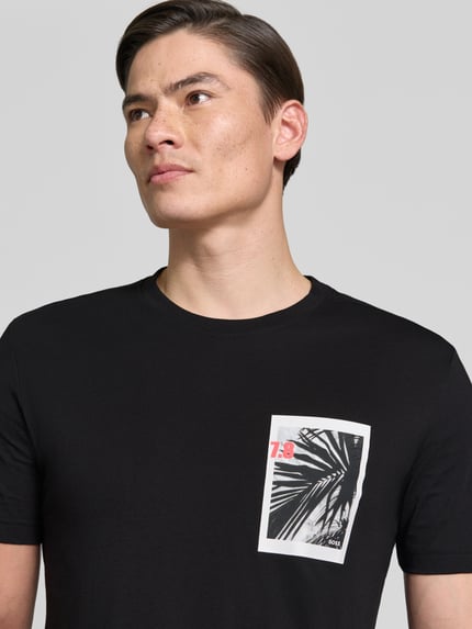Męski model nosi Regular Fit T-Shirt aus reiner Baumwolle Modell 'TE_BOARD' von BOSS Orange w kolorze czarnym, krój regular fit, okrągły dekolt, krótki rękaw, nadruk foto i aplikacja label, dzianina jersey z czystej bawełny, zbliżenie na górną część ciała.