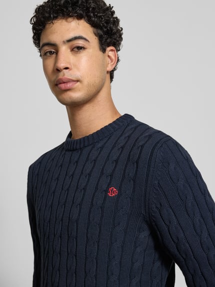 Männermodel trägt Slim Fit Pullover in Strick-Optik von Superdry aus reiner Baumwolle, Logo-Stitching, Rundhalsausschnitt, Nahaufnahme Oberkörper.