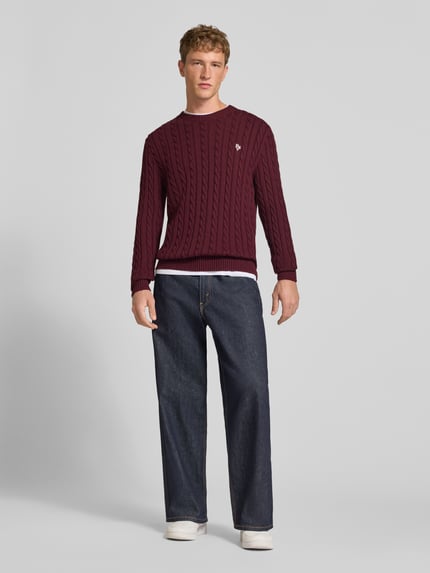 Männermodel trägt Slim Fit Strickpullover aus reiner Baumwolle mit Zopfmuster von REVIEW, bordeauxfarben, Label-Stitching, Jeans, Ganzkörperansicht von vorne.