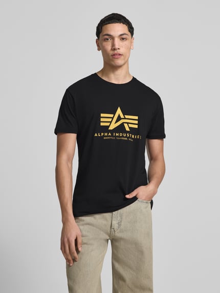 Männermodel trägt T-Shirt mit Label-Detail Modell 'Basic' von Alpha Industries aus Reine Baumwolle, Gerader-Schnitt, Rundhalsausschnitt, schwarze Farbe mit gelbem Aufdruck, Rückenlänge bei Größe M: 67 cm, Ganzkörperansicht von vorne.