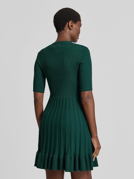 Frauenmodel trägt Strickkleid in Minilänge mit Viskose-Anteil von Christian Berg Woman in Dunkelgrün, Ripp-Optik, Rüschensaum, taillierte Passform, kurzärmelig, Ganzkörperansicht Rückansicht.