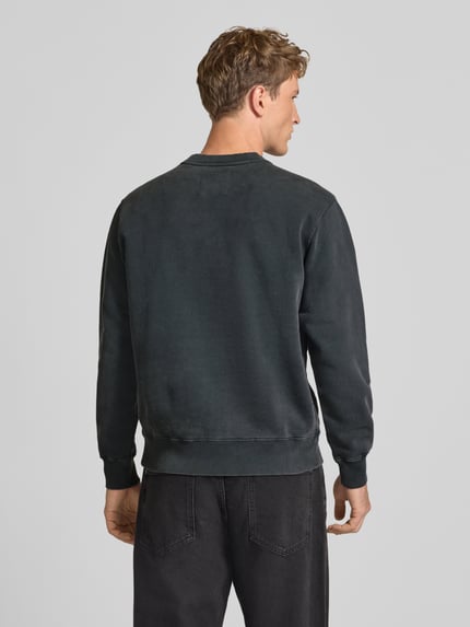 Männermodel trägt Sweatshirt aus reiner Baumwolle mit Stickerei Modell 'Axiom' von Edwin in Dunkelgrau mit gerippten Abschlüssen, Ganzkörperansicht Rückansicht. Relaxed Fit, langärmliges Design, überschnittene Schultern, Rückenlänge Größe M 65 cm, Ärmellänge 62 cm.