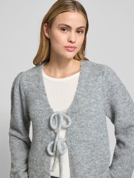 Kobiece model prezentuje szarą swetrową Strickjacke mit Schnürverschluss Modell 'GRSLULU' od FREE/QUENT, mieszanka wełny, luźny krój, prążkowane wykończenia, zapięcie na sznurek, dekolt V, długość tyłu 50 cm, długość rękawa 64 cm; widok na górną część ciała z przodu.