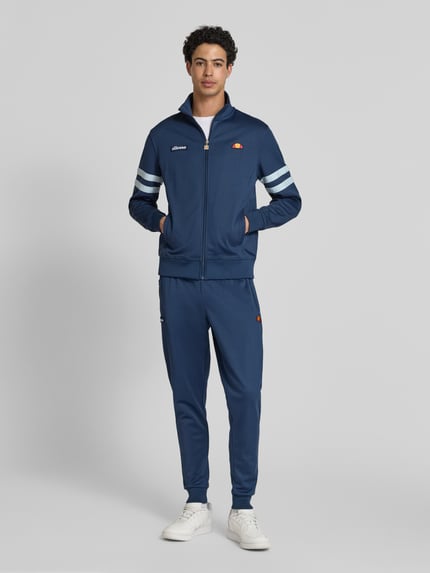 Männermodel trägt Sweatjacke aus Baumwoll-Mix Modell 'ROMA' von Ellesse in Blau mit Label-Details, Gerader Passform, Stehkragen, Reißverschluss, Eingrifftaschen, elastischen Abschlüssen, Ganzkörperansicht von vorne.