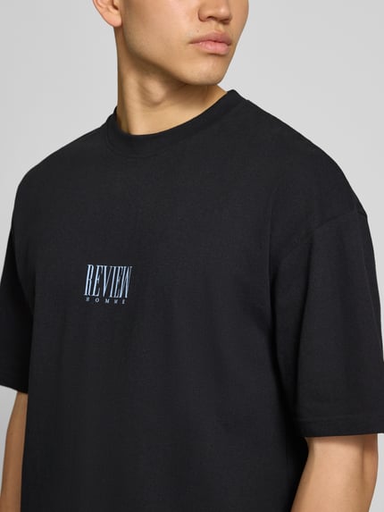 Männermodel trägt T-Shirt mit Logo-Print und Rundhalsausschnitt von REVIEW, schwarze Reine Baumwolle, Loose-Fit, Nahaufnahme Oberkörper.