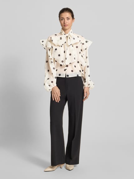 Frauenmodel trägt Regular Fit Schluppenbluse mit Rüschen Modell 'DOROTHY' von Vero Moda, Optik:Allover-Muster, Semitransparent, Polka-Dots, Kragenform: Mock Neck mit Schluppe, Applikation: Rüschen, Verschluss: einreihige Knopfleiste, Ärmelform: Langarm mit Ballonärmel, Ärmelabschluss: Elastisch, Ganzkörperansicht von vorne.
