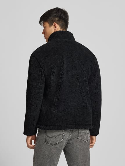 Männermodel trägt Fleecejacke mit Label-Detail und Reißverschluss von REVIEW in Schwarz, Teddyfell-Struktur, Ganzkörperansicht Rückansicht. Regular Fit, Stehkragen, Rückenlänge bei Größe M: 70 cm, Schulterbreite: 56 cm, Brustweite: 59 cm, Ärmellänge: 64 cm.