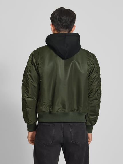 Männermodel trägt Bomberjacke mit Kapuze und Reißverschlusstaschen von Superdry in Olivgrün, Regular-Fit, Rückenansicht, Kontext: Herren Bomberjacke von Superdry, Kapuze, Reißverschluss, Reißverschlusstaschen, Rückenlänge 56 cm, Schulterbreite 45 cm, Brustweite 60 cm, Ärmellänge 68 cm. Ganzkörperansicht Rückansicht.