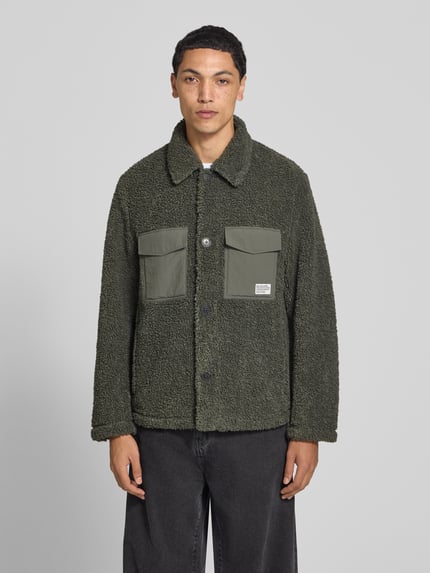Męski model nosi Regular Fit Hemdjacke mit Brustleistentaschen od Marc O'Polo DENIM w kolorze ciemnozielonym, faktura pluszowa, guziki, dwie kieszenie na piersi, widok całej sylwetki z przodu.