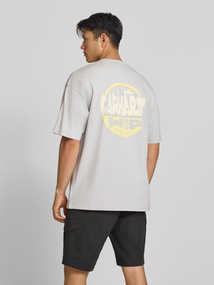 Männermodel trägt Relaxed Fit T-Shirt aus reiner Baumwolle Modell 'Luggage' von Carhartt Work In Progress in Beige mit großem gelbem Rückenprint, lockere Passform, schwarze Shorts, ganzkörperansicht Rückansicht, Herren T-Shirt Relaxed Fit aus reiner Baumwolle, Label-Detail, Rückenlänge Größe M 78 cm.