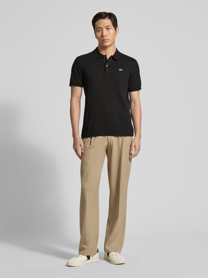 Mannelijk model draagt Regular Fit Poloshirt uit zuivere katoen van Lacoste, effen zwart, polokraag, korte knoopsluiting, logo detail, regular fit, ribgebreide korte mouwen, asymmetrische zoom met zijsplitten, materiaal zuivere katoen. Volledige lichaamsaanzicht van voren.