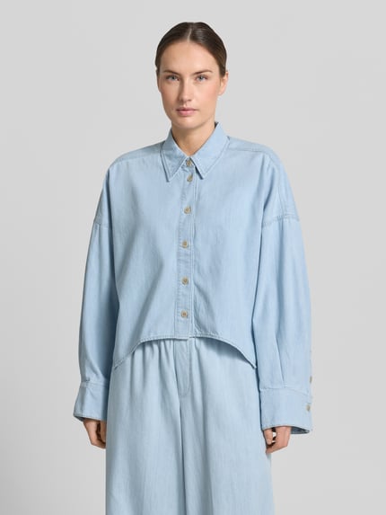 Frauenmodel trägt Relaxed-Fit Damen Hemdbluse in Denim-Optik Modell 'Oshia' von Drykorn aus Reine Baumwolle, Knopfleiste, Langärmelig, Hemdblusenkragen, hellblaue Farbe, glatte Textur, Rückenlänge 55 cm, Ärmellänge 47 cm, Ganzkörperansicht von vorne.