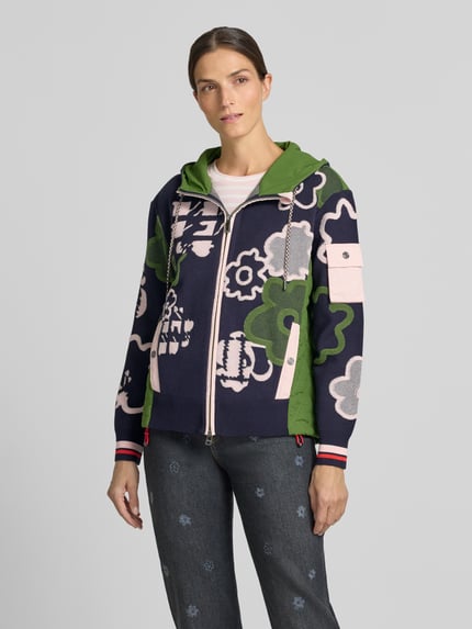 Frauenmodel trägt Regular Fit Jacke mit floralem Allover-Stitching von Marc Cain in Dunkelblau, Grün und Rosé mit Kapuze, Eingrifftaschen, elastischen Ärmelabschlüssen und Zweiwege-Reißverschluss, Ganzkörperansicht von vorne.