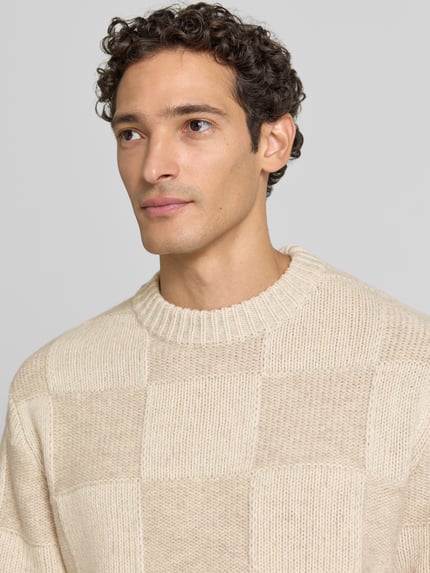 Męskimm model ma na sobie sweter z dzianiny z okrągłym, prążkowanym dekoltem Strickpullover mit geripptem Rundhalsausschnitt von Les Deux, bawełna, prosty krój, prążkowane wykończenia, długość tyłu w rozmiarze M: 72 cm, długość rękawów w rozmiarze M: 66 cm, kolor beżowy w kratę, faktura wyraźnie widoczna, zbliżenie na górną część tułowia