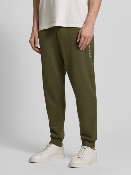 Męski model nosi Comfort Fit Sweatpants aus Baumwoll-Mix od Tommy Hilfiger, oliwkowe, materiał sweat, jednokolorowy, aplikacja logo, ściągacze, kieszenie, widok z przodu, zbliżenie na dolną część ciała.