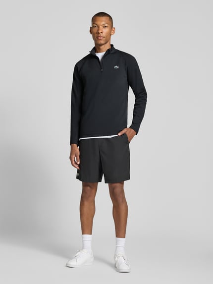 Męskki model nosi Regular Fit Sport-Langarmshirt mit Ultra-Dry-Technologie od Lacoste w kolorach czarnym i białym, sportowa faktura tkaniny, Ganzkörperansicht von vorne.