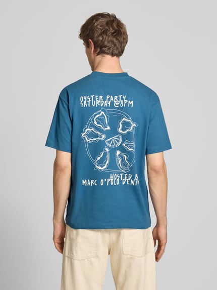 Männermodel trägt Relaxed Fit T-Shirt aus reiner Baumwolle mit Motiv-Print von Marc O'Polo Denim, Optik: Unifarben, Passform: Relaxed-Fit, Motiv-Print, Rundhals, Ärmelform: Kurzarm, Material: reine Baumwolle, kräftiges Blau mit weißem Print. Ganzkörperansicht Rückansicht.