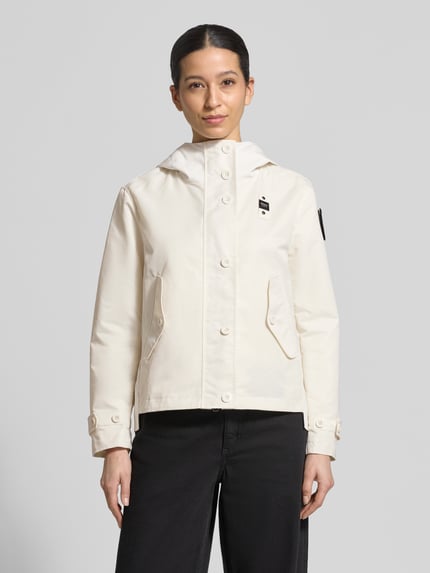 Modelka nosi Jacke mit Label-Patches Modell 'MARCELA' od Blauer Usa, prosta forma, kaptur, naszywki, beżowy kolor, gładka faktura, patki z guzikami, długość tyłu w rozmiarze S: 61 cm, szerokość ramion w rozmiarze S: 44 cm, szerokość w rozmiarze S: 53 cm, długość rękawa w rozmiarze S: 59 cm. Widok całej sylwetki od przodu.