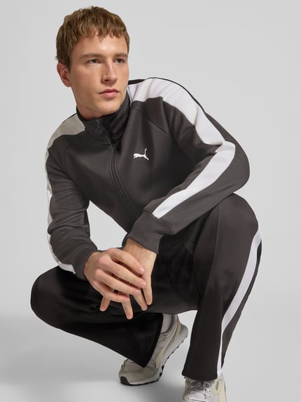 Mężczyzna nosi Sweatjacke mit Label-Print od PUMA PERFORMANCE, czarny materiał z białymi pasami, krój regular fit, stehkraag, zamek, ściągacze, kieszenie na zamek, mieszanka bawełny, widok całej sylwetki z przodu.