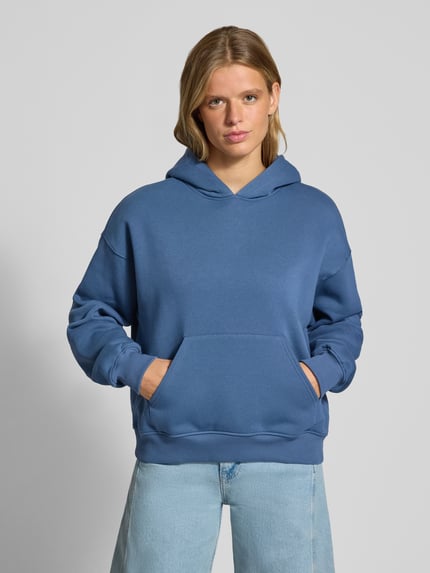 Frauenmodel trägt Hoodie mit Känguru-Tasche von Gina Tricot in Blau, Baumwoll-Mix, Relaxed Fit, überschnittene Schultern, Ganzkörperansicht von vorne.