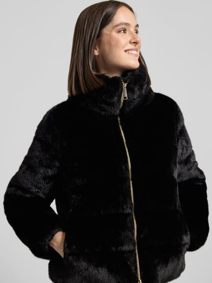 Frauenmodel trägt schwarze, glänzende Webpelzjacke mit Zweiwege Reißverschluss von S. OLIVER BLACK LABEL, Struktur, seitliche Taschen, Ganzkörperansicht von vorne.