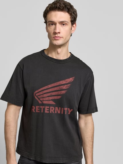 Mężczyzna nosi T-Shirt z nadrukiem label-Print model 'AÉRO' od RETERNITY, czarna bawełna, krój regular fit, okrągły dekolt, długość tyłu w rozmiarze M: 67 cm, zdjęcie całej sylwetki z przodu.