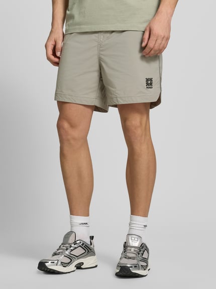 Mężczyzna nosi Regular Fit Shorts mit Label-Applikation Modell 'COLAS262S' od HUGO, kolor jednolity, kontrastowe paski, elastyczny pas, kieszenie, zbliżenie na nogi