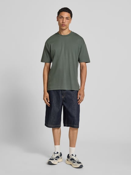 Mannelijk model draagt Relaxed Fit T-shirt uit zuivere katoen van Marc O'Polo Denim in olijfgroen met rechte zoom, gecombineerd met donkere denim shorts; volledige lichaamsaanzicht van voren.