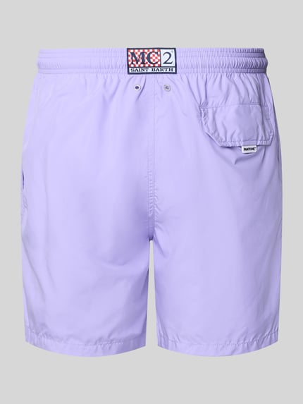 Männermodel trägt Badehose mit Logo-Print Modell 'Lighting Pantone' von MC2 Saint Barth in Lila mit Logo-Patch, Ganzkörperansicht Rückansicht.
