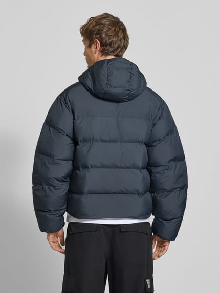 Männermodel trägt Regular Fit Puffer Jacket mit Kapuze von adidas Originals, jednokolorowy, hydrofobowy, krój regular fit, kieszenie zapinane na zamek błyskawiczny, detal z logo, długi rękaw, elastyczne zakończenia, wykończenie materiału: tkanina, Ganzkörperansicht Rückansicht.